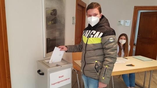 Junger Wähler mit Corona-Maske vor einer Wahlurne mit Stimmzettel in der Hand